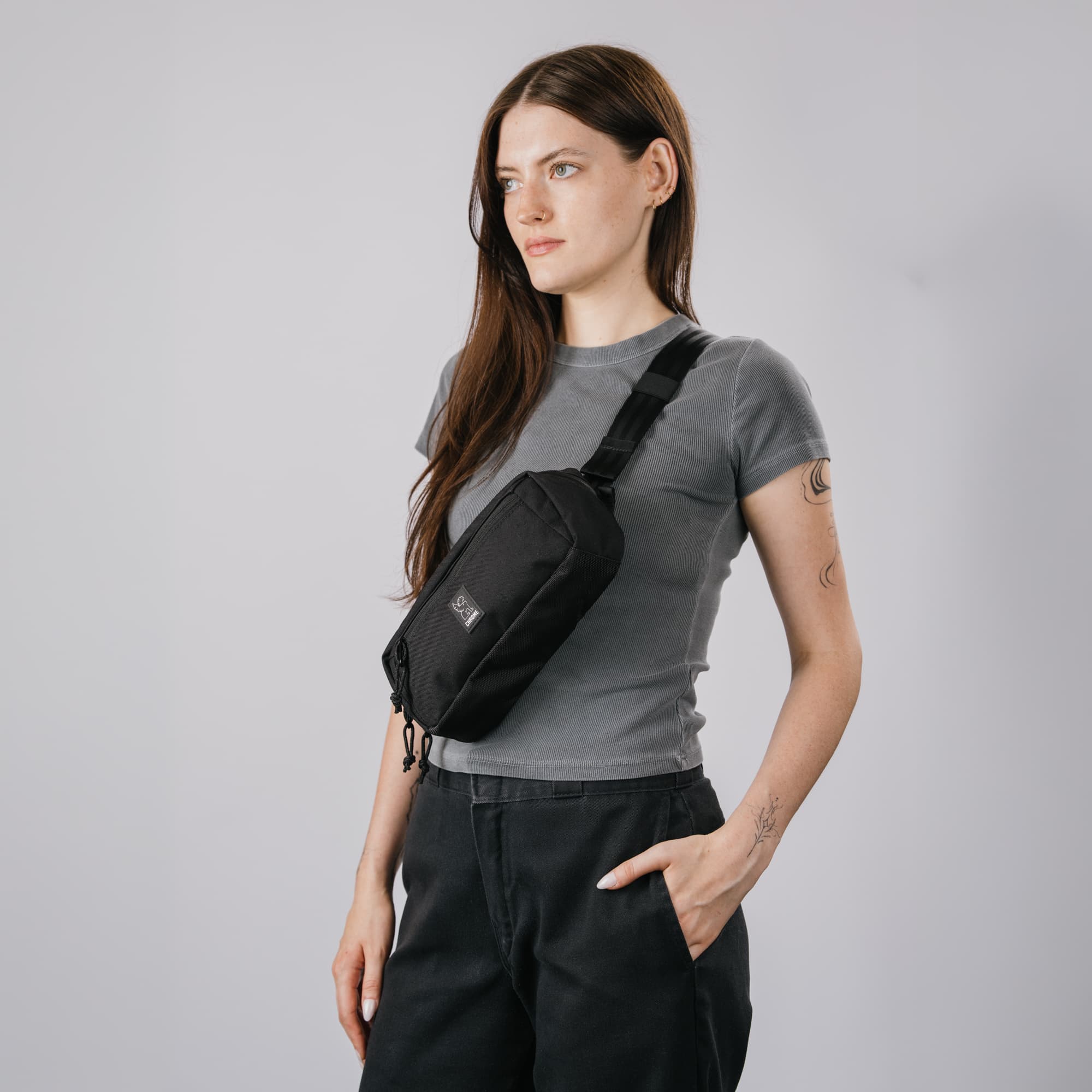 A woman wearing a gray t-shirt and black pants models a black Micro Kadet crossbody sling bag with a Chrome logo patch on the front, set against a plain light gray studio background.