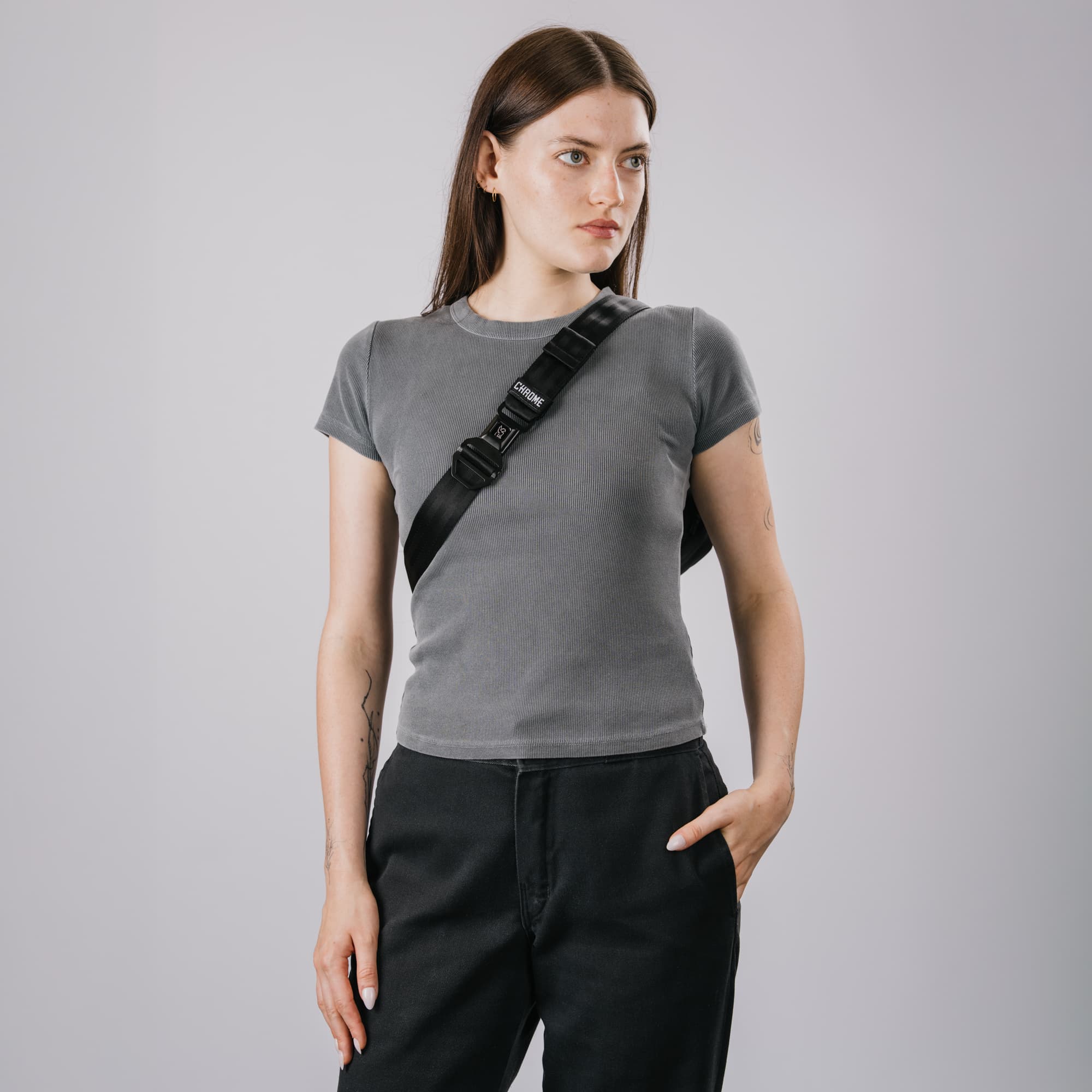 A woman modeling a dark gray short-sleeve crewneck t-shirt with a black adjustable crossbody Micro Kadet sling bag worn across her chest, paired with black pants, against a solid light gray studio background.