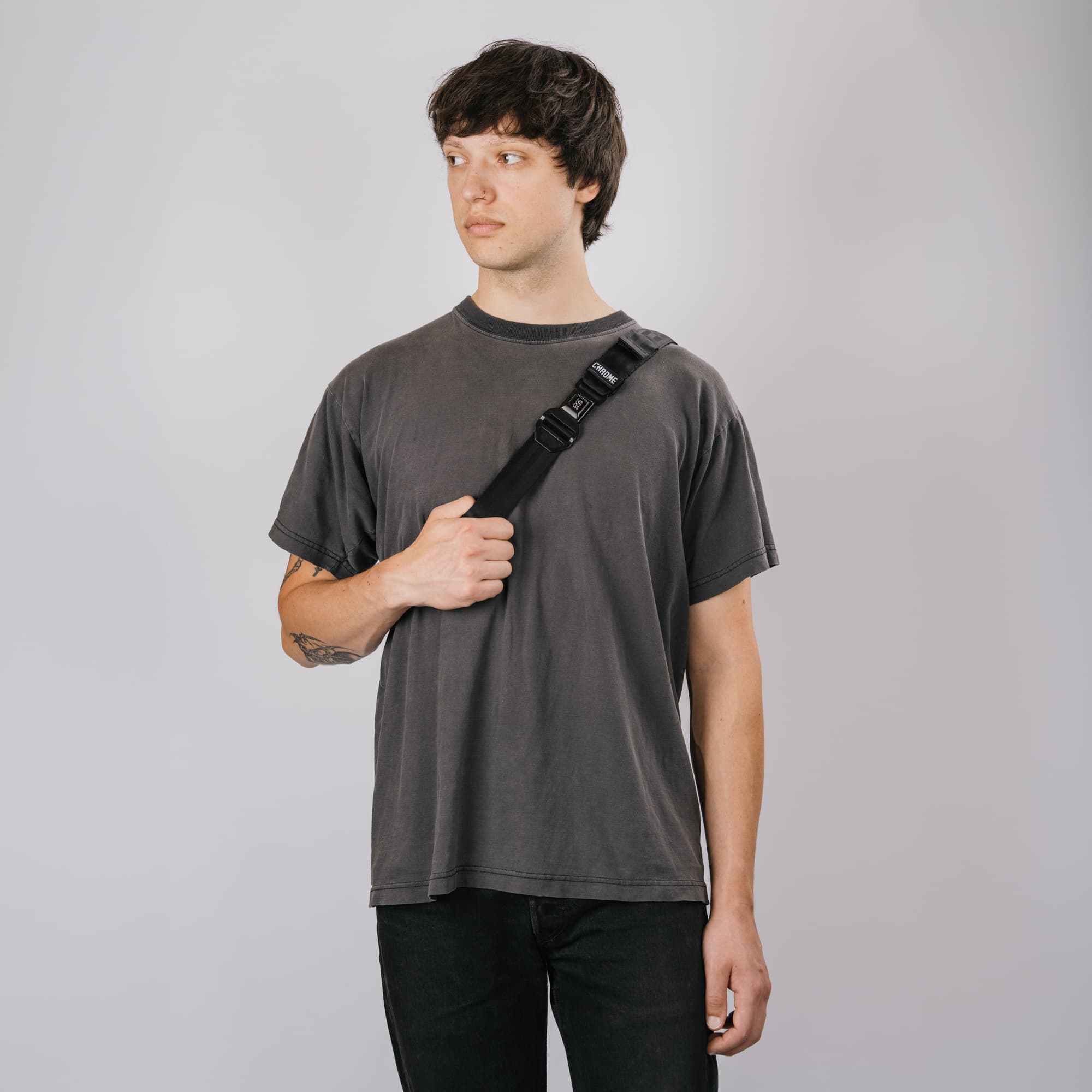 A man wearing a washed charcoal gray t-shirt and black jeans, carrying a black Micro Kadet crossbody bag across his chest, set against a plain light gray studio background.