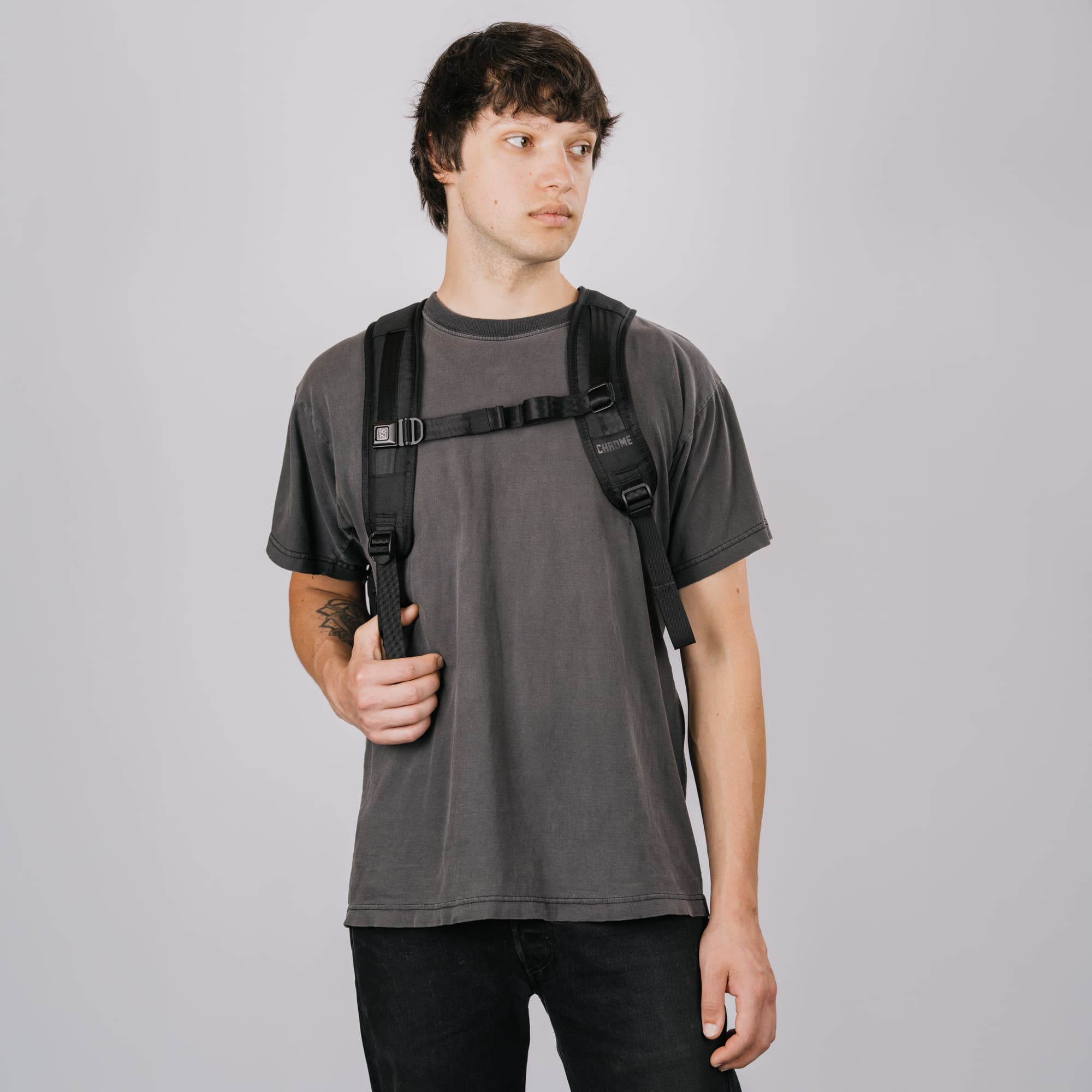 A man wearing a dark gray t-shirt and black pants, carrying a new black Recruit 22L backpack with shoulder straps over his shoulders, against a solid gray studio background.