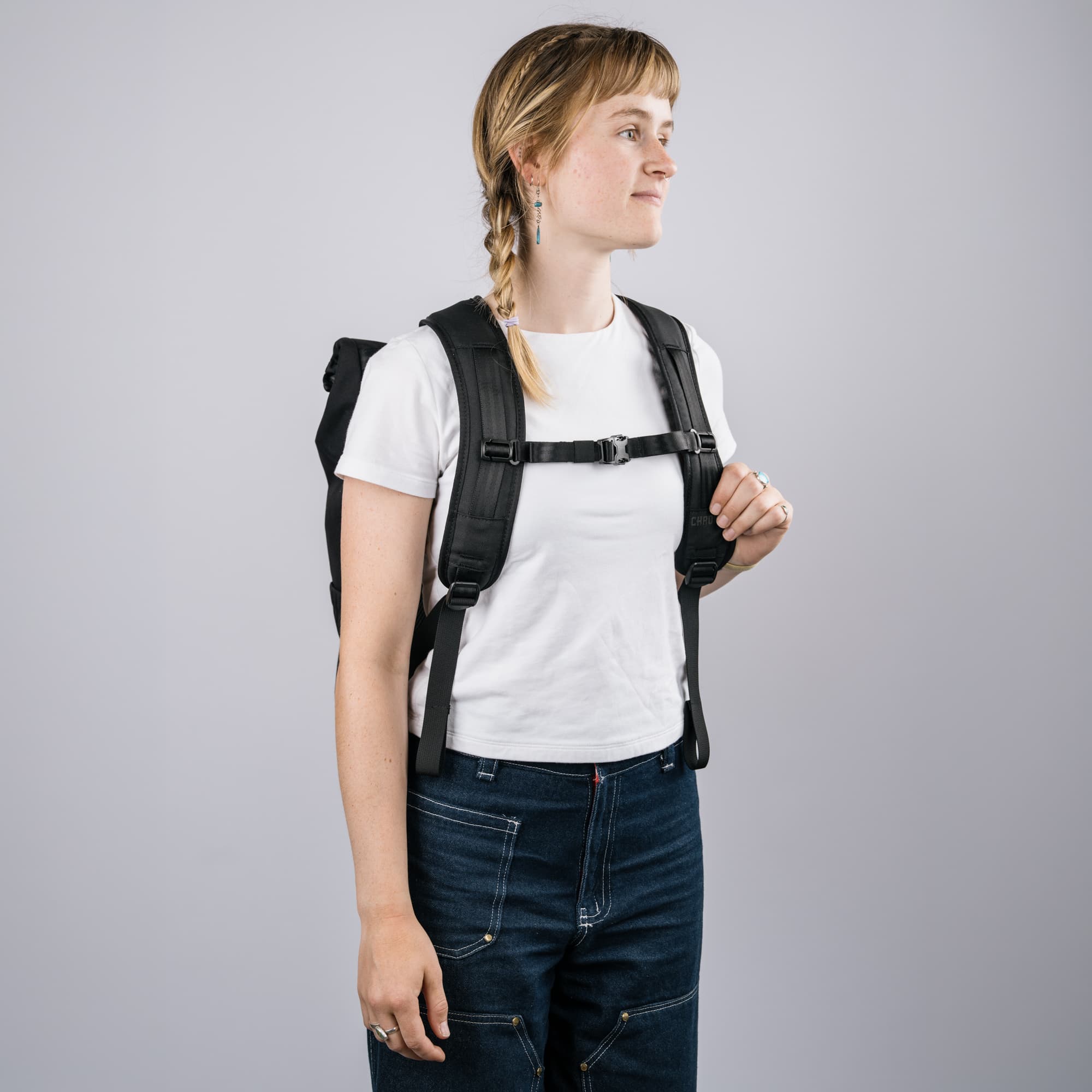 A woman wearing a white t-shirt and dark blue jeans models the Black Initiate Pack with a chest harness strap system against a solid gray background.