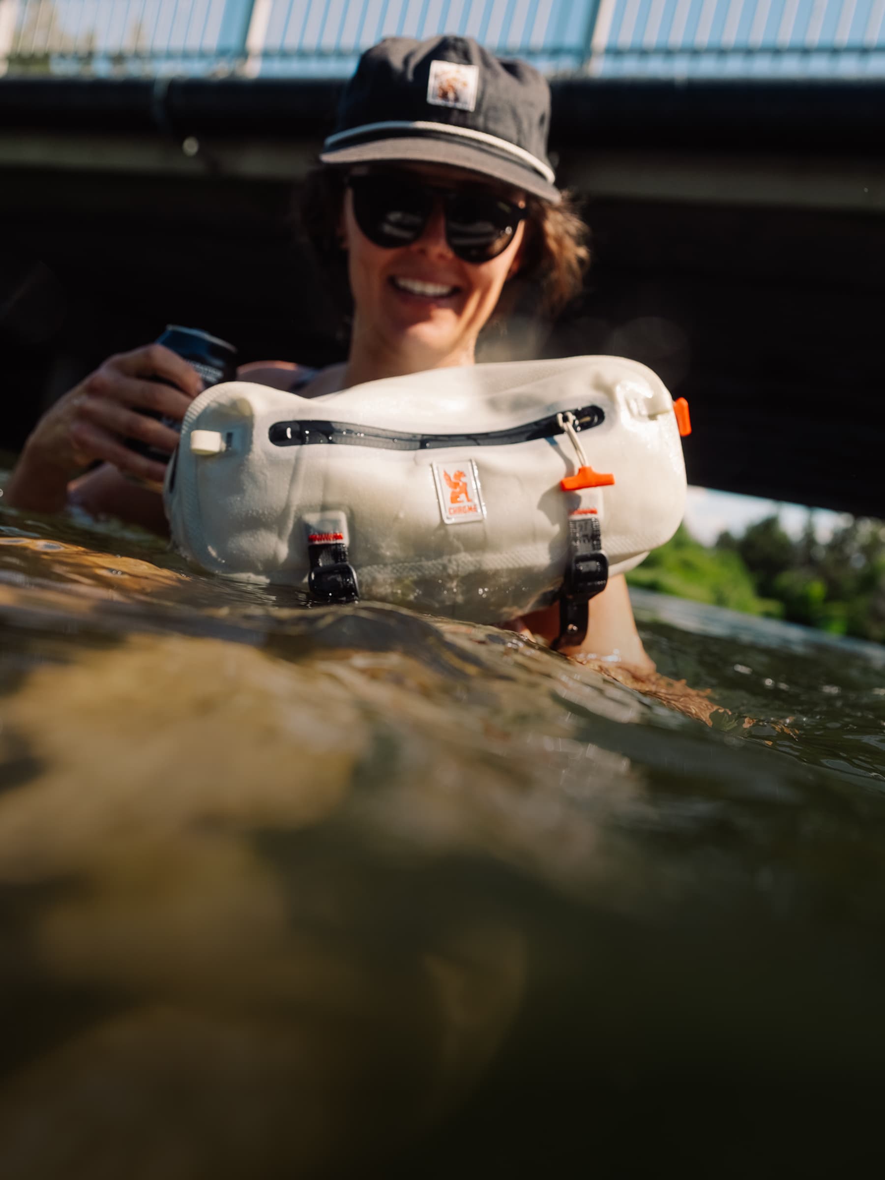 A person in shallow water wearing a black cap and sunglasses holds a Mini Kadet waterproof dry bag with black straps and orange accents. The photo is taken outdoors under a bridge with water and greenery in the background.
