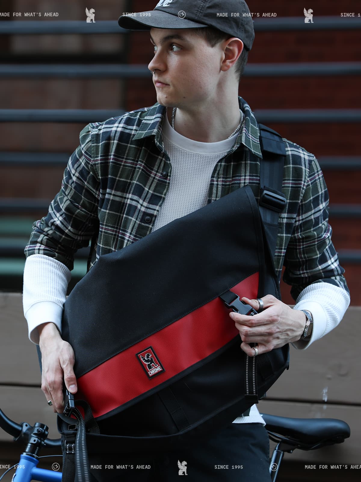 Person holding a black and red bag with a brand logo, wearing a plaid shirt and cap.