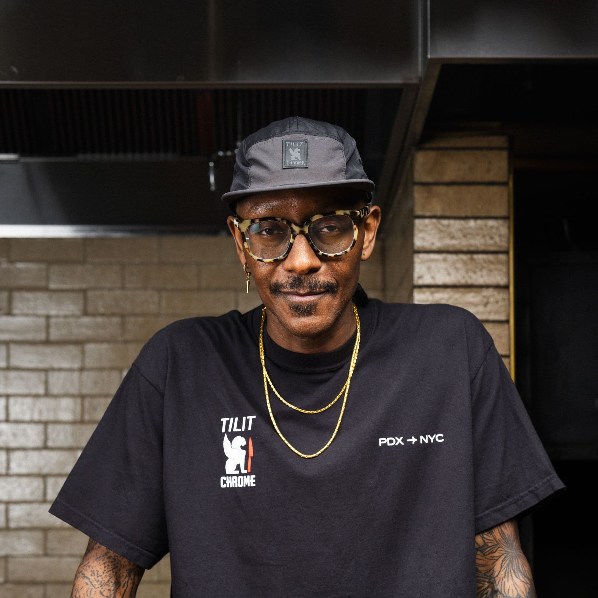 Gregory Gourdet wearing the commuter cap with visible Chrome x TILIT branding, standing against a brick wall.