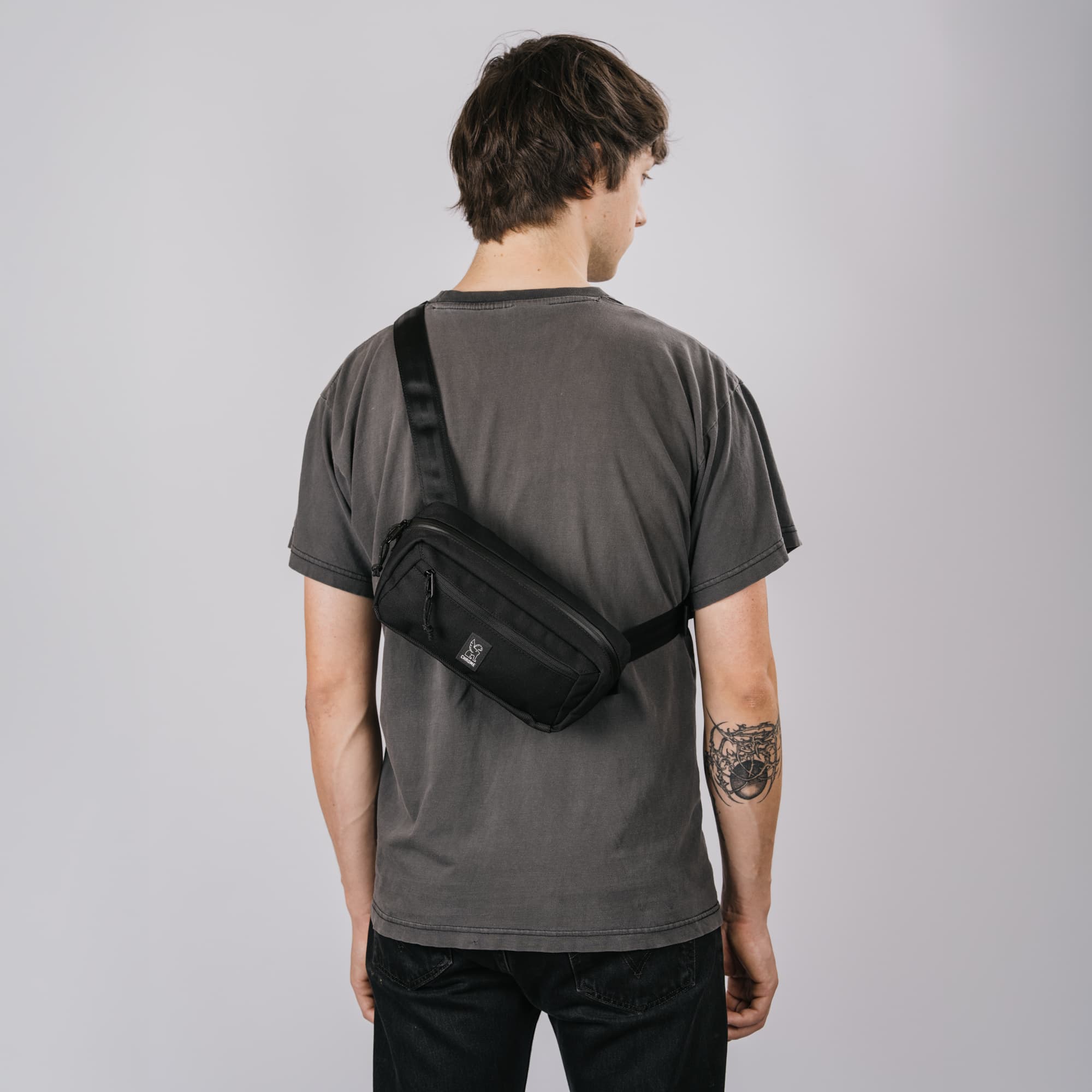 A man viewed from behind wearing a black crossbody Micro Kadet sling bag across his back, paired with a faded dark gray t-shirt and black jeans, against a solid light gray studio background.