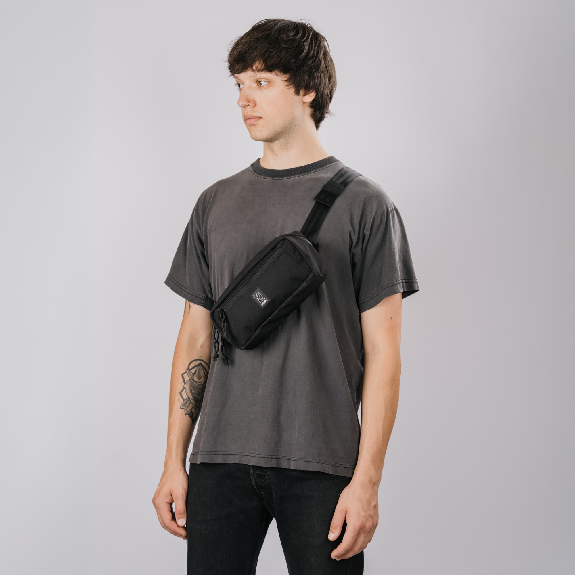 A man wearing a black Micro Kadet 3L crossbody sling bag across his chest, styled with a faded dark gray t-shirt and black jeans, photographed against a solid light gray studio background.