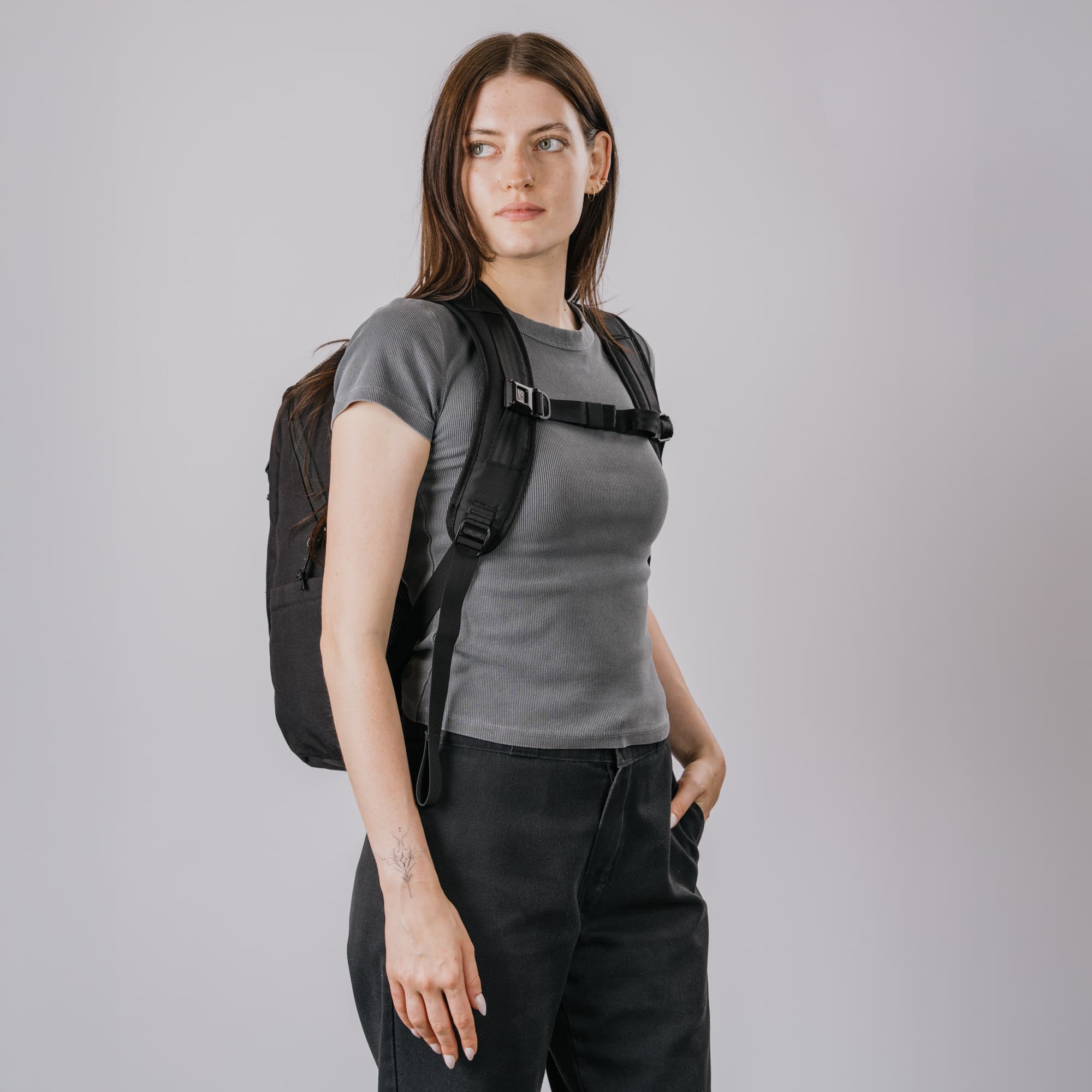 A woman wearing a gray t-shirt and black pants, carrying a black Recruit 22L backpack with a chest strap over her shoulders, set against a solid light gray background.