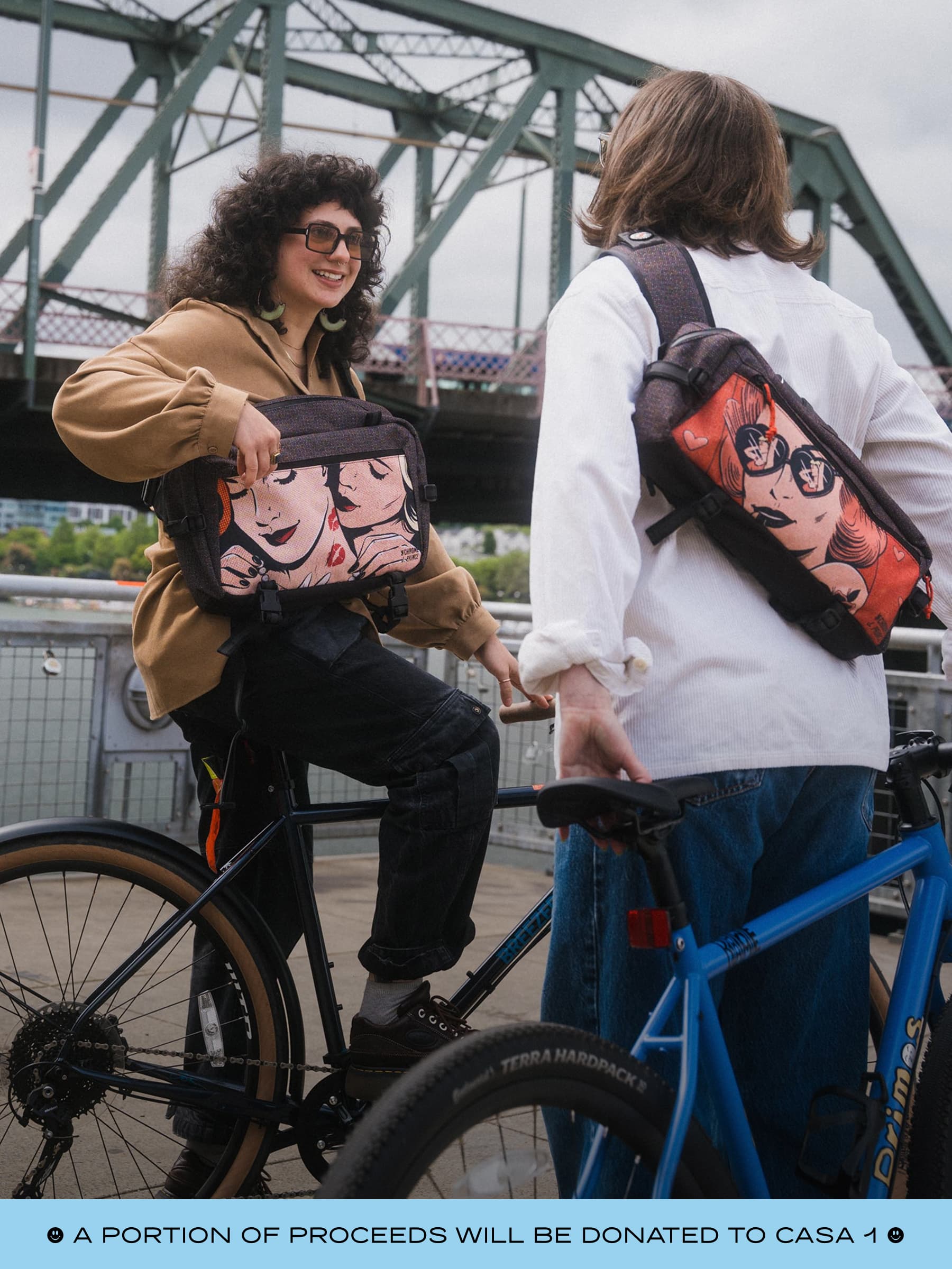 Two people with the Pride print Kadets standing next to bicycles, with a bridge in the background.